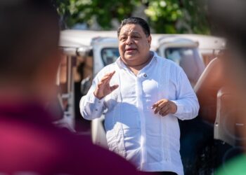 Hombre con guayabera blanca habla al aire libre frente a vehículos blancos