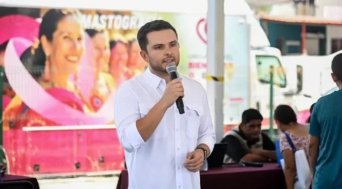 Hombre con camisa blanca habla con micrófono frente a una unidad móvil de mastografía