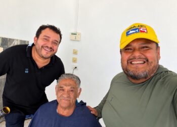 Tres hombres sonríen para una foto; uno mayor, sentado y cubierto con una capa azul, al centro