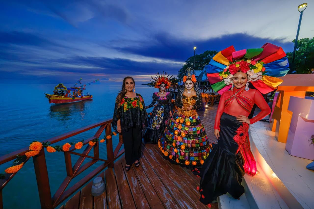 Mara Lezama junto a catrinas en el muelle de Calderitas, con una trajinera en la Bahía de Chetumal
