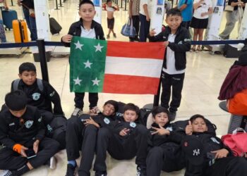 Niños de la Selección B de futbol de Yucatán, con ropa deportiva negra, esperan sentados en el piso del aeropuerto de Cancún junto a la bandera de Yucatán