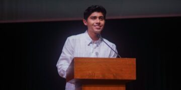 Joven con guayabera blanca habla desde un atril de madera en la ceremonia de graduación de la UQROO Campus Playa del Carmen